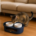 Load image into Gallery viewer, Tabby cat approaching a navy blue elevated dual pet food bowl feeder with white ceramic bowls and storage compartment on wooden floor near beige sofa.
