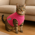 Load image into Gallery viewer, Tabby cat wearing a pink fleece vest standing on a wooden floor in front of a beige couch.
