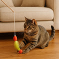 Load image into Gallery viewer, Tabby cat playing with red and yellow feather wand toy on wooden floor near beige sofa.

