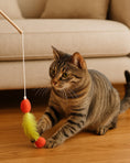 Load image into Gallery viewer, Tabby cat playing with red and yellow feather wand toy on wooden floor near beige sofa.
